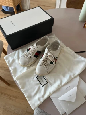 Gucci sneakers  - Snygga vita sneakers från Gucci med klassiska röd-gröna ränder på sidorna och broderad detalj. Skorna är i läder och har en låg siluett. Perfekta för dig som vill ha en lyxig och trendig look.