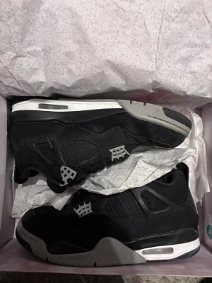 Svarta Air Jordan 4 Retro sneakers - Snygga Air Jordan 4 Retro sneakers i svart med grå och vita detaljer. Skorna har en klassisk siluett med meshpaneler, plastdetaljer och synlig Air-dämpning i sulan. Perfekta för dig som gillar streetwear och vill ha en ikonisk look.