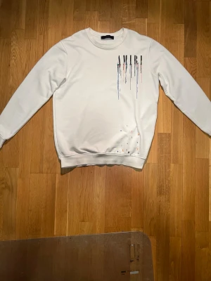 Vit sweatshirt från Amiri - Vit sweatshirt från Amiri med färgstänk och droppande logotyptryck både fram och bak. Tröjan har rund halsringning, långa ärmar och ribbade muddar. Perfekt för dig som gillar streetwear med unik design.