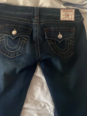 Mörkblå True Religion jeans - Snygga mörkblå jeans från True Religion med ikoniska kontrastsömmar och stora fickor bak med dekorativa knappar. Klassisk femficksmodell och silverfärgade detaljer. Perfekta för dig som gillar streetwear och vill sticka ut med coola detaljer.