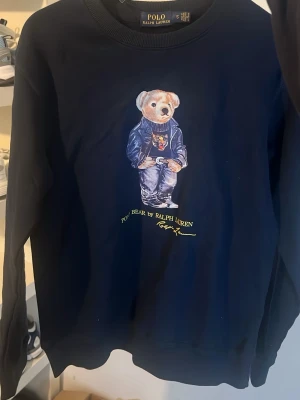 Ralph lauren tröja - Snygg mörkblå tröja från Ralph Lauren med ikoniska Polo Bear-trycket framtill. Tröjan har rund halsringning, långa ärmar och ribbade muddar. Tillverkad i mjuk bomull som är skön att ha på sig. Perfekt för dig som gillar streetstyle och klassiska märken.