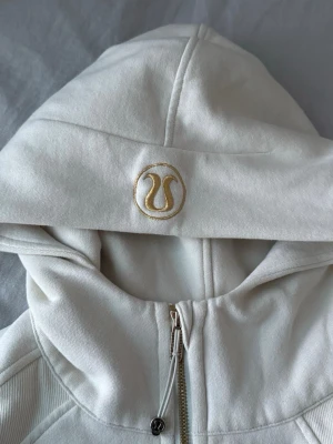 Vit hoodie från Lululemon med dragkedja - Snygg vit hoodie från Lululemon med halv dragkedja framtill och broderad logga i guld på huvan. Hoodien har känguruficka, ribbade muddar och är tillverkad i mjukt, tjockt material som känns riktigt lyxigt. Oanvänd. (Köpt i USA så har ingey kvitto men den är äkta). Tröjan är oversized på mig som vanligtvis har på S/M.