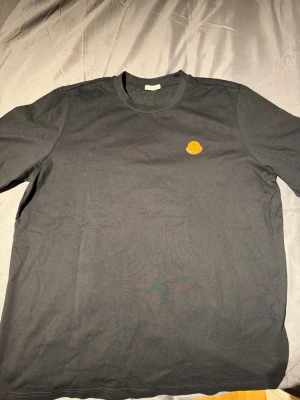 Svart Moncler t-shirt med logga - Snygg svart t-shirt från Moncler Moncler-logga på bröstet i läder. Tillverkad i mjuk bomull och har en enkel, stilren design som passar till det mesta. Perfekt för dig som gillar exklusiva märken och clean stil. Frågor? Bara att skriva. Mvh Noah
