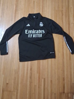 Svart Real Madrid träningströja Adidas - Säljer en svart långärmad träningströja från Adidas med Real Madrid-logga och Emirates Fly Better-tryck på bröstet. Tröjan har vita detaljer och dragkedja vid halsen. Perfekt för fotbollsträning eller sportiga dagar.
