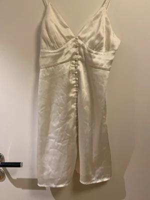 Vit satin klänning med knappar, BIANCA x Nelly.com - Säljer en elegant vit klänning i satin med smala axelband och dekorativa knappar framtill. Klänningen har en figurnära passform upptill och lätt utställd kjol. 