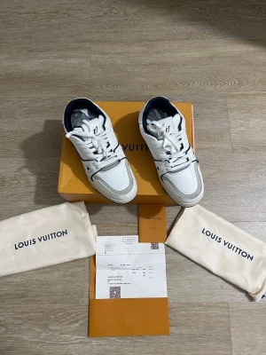  vita sneakers - Snygga vita sneakers med marinblå detaljer och perforerade partier. Klassisk LV-logga på sidan och signaturdetaljer på sulan. Skorna har en chunky sula och är tillverkade i läder med textilfoder. Perfekta för dig som vill ha en exklusiv och trendig look.