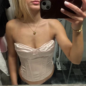 Ljusrosa satin korsett med bygel - Säljer en ljusrosa korsett i glansigt satinmaterial med bygel och markerade sömmar. Axelbandslös modell med formade kupor och bred rygg. Perfekt för att skapa en snygg siluett och ge stöd.