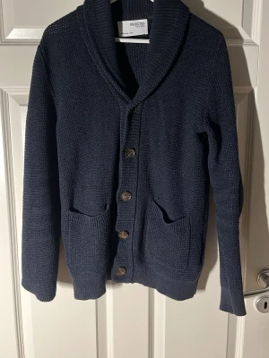Mörkblå stickad kofta från Selected Homme - Säljer en mörkblå stickad kofta från Selected Homme i storlek M. Koftan har grovstickad struktur, bred krage, 5 knappar framtill och två praktiska fickor. Perfekt för lager-på-lager och chill stil under kyliga dagar. Vid frågor bara skriv// Almen