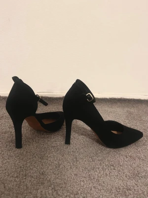 Svarta pumps med hög klack och rem - Säljer ett par eleganta svarta pumps med spetsig tå och hög klack. Skorna har en smal rem med guldfärgat spänne runt ankeln och är tillverkade i mjuk mocka. Perfekta för att lyfta vilken outfit som helst.