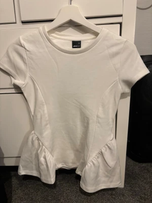 Vit peplum topp från Gina Tricot - Säljer en vit peplum topp från Gina Tricot i storlek XS. Toppen har rund hals, korta ärmar och en snygg volangdetalj nedtill som ger en feminin siluett. Materialet är mjukt och känns bekvämt mot huden. Perfekt att styla med jeans eller kjol.