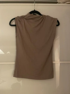 Beige ärmlös polotopp - Säljer en beige polotopp med ärmlös design. Toppen har en mjuk och slät yta, perfekt för att styla med jeans eller kjol. Enkel och stilren look med hög hals som ger en cool vibe. Passar dig som gillar minimalistiska plagg.