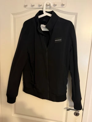 Svart jacka från Calvin Klein Jeans - Svart bomberjacka från Calvin Klein Jeans med stilren design och logotyp på bröstet. Jackan har dragkedja framtill, ribbade muddar vid ärmslut och nederkant samt ståkrage. Perfekt för dig som gillar enkel och snygg streetstil.