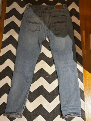 Ljusblå Nudie Jeans - Säljer ett par ljusblå jeans från Nudie Jeans med klassisk femficksdesign och snygga broderade detaljer på bakfickorna. Jeansen är tillverkade i mjukt bomullsmaterial med lite stretch för extra komfort. Perfekt för dig som gillar en smal siluett.