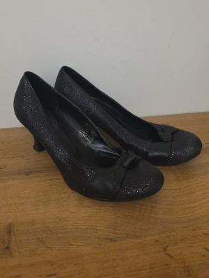 Svarta glittriga pumps med rosett - Säljer ett par svarta pumps med glittrigt tyg och en söt rosett framtill. Skorna har rund tå och låg klack, vilket gör dem både stilrena och trendiga. Perfekta för dig som vill ha lite extra bling på fötterna. Materialet är syntet med glittrig finish.