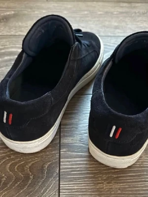 Svarta sneakers i mocka med snörning - Säljer ett par svarta sneakers med vit sula och snörning. Skorna är gjorda i mocka och har en stilren design med små broderade detaljer i rött och vitt på hälen. Perfekta för dig som gillar klassisk streetstil.