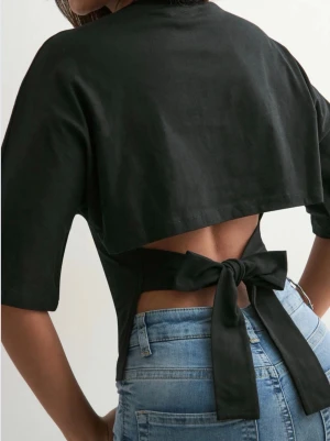 Svart croppad topp med knyt i ryggen - Säljer en svart croppad topp med korta ärmar och snygg öppen rygg som knyts med en stor rosett baktill. Toppen har en enkel, stilren framsida och är gjord i mjuk bomull. Perfekt för dig som vill ha en cool och trendig look.