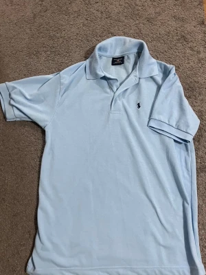 Polo Ralph Lauren piké - Snygg ljusblå pikétröja från Polo Ralph Lauren med klassisk krage och korta ärmar. Tröjan har den ikoniska broderade loggan på bröstet och är tillverkad i mjuk bomull. Perfekt för en avslappnad men stilren look. L/M