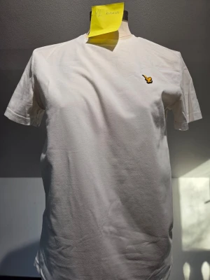 Lyle scott t-shirt - Klassisk vit t-shirt från Lyle & Scott med gul fågellogga broderad på bröstet. Enkel och stilren design med rund hals och korta ärmar. Perfekt till jeans eller shorts för en clean look.