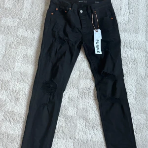 Svarta slim fit jeans från Purple Brand - Svarta jeans från Purple Brand i storlek 32 med slim fit passform. Byxorna har slitna detaljer på framsidan och klassisk femficksdesign. Materialet är jeans och de har knappstängning framtill. Snygga att matcha med sneakers och hoodie.