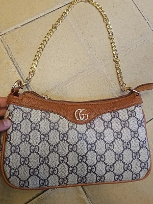 handväska med kedja och GG-mönster - Säljer en snygg handväska med klassiskt GG-mönster i beige och brunt. Väskan har detaljer i brunt skinn, guldfärgad kedja och dragkedja upptill. Perfekt storlek för det viktigaste och har en lyxig känsla med ikonisk logga framtill.