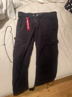 Svarta cargopants från Alpha Industries - Svarta cargopants från Alpha Industries med flera stora fickor på benen och klassisk logopatch bak. Byxorna har normal passform, dragkedja och knapp i midjan samt detaljer som snörning vid benslut. Materialet är slitstark bomull, perfekt för dig som gillar utility-vibe.