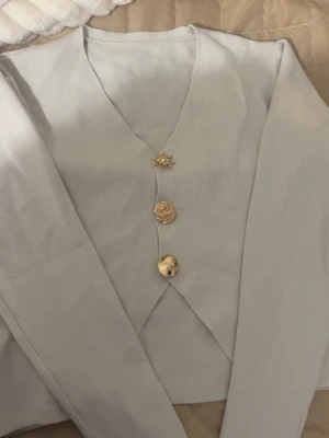 Beige kofta med guldknappar - Snygg beige kofta med unik design och tre dekorativa guldknappar framtill. Koftan har v-ringad hals, långärmade ärmar och en lätt croppad passform med slits framtill. Perfekt för dig som gillar stilrena detaljer. Den är oanvänd