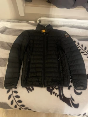 Svart dunjacka från Parajumpers - Svart dunjacka från Parajumpers med quiltad design och hög krage. Jackan har dragkedja framtill, två sidofickor med dragkedja och en gul detalj vid.