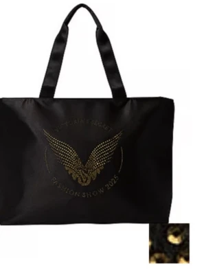 Svart shoppingväska Victoria's Secret - Stor svart shoppingväska från Victoria's Secret med Fashion Show 2025-tryck i guldiga nitar och ikoniska änglavingar på framsidan. Väskan har breda handtag och är gjord i slitstark polyester, perfekt för att bära med sig allt man behöver.