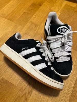 Adidas Campus sneakers svart/vit strl 38 - Säljer ett par Adidas Campus sneakers i svart mocka med vita detaljer och klassiska tre ränder på sidan. Skorna har tjocka vita snören, gummisula i brun färg och rund tå. Adidas-logga på plösen och insidan. Perfekt för dig som gillar retrostil och streetwear.