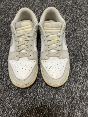 Nike Dunk Low i ljusgrå och vit - Nike Dunk Low sneakers i ljusgrått och vitt med klassisk swoosh på sidan. Skorna har snörning, perforerad tå och platt sula. Ovandelen är i skinn och syntet, med broderad Nike-logga på hälen. Perfekt för dig som gillar clean och stilren look.