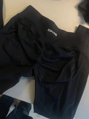 Svarta leggings från DFYNE - Säljer ett par svarta leggings från DFYNE med bred ribbad midja och snygg scrunch-detalj bak. De är tillverkade i ett stretchigt syntetmaterial som sitter tight och är perfekta för träning. Loggan är tryckt framtill på midjan.