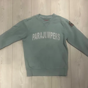 Grön fleece tröja från Parajumpers - Säljer en grön tröja från Parajumpers med broderad logga på ärmen och text på bröstet. Tröjan har långa ärmar och ribbade muddar vid ärmslut och nederkant. Perfekt tröja