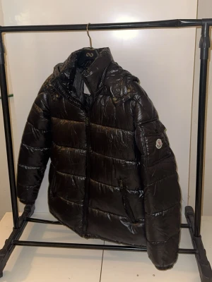 Svart pufferjacka från Moncler - Svart glansig pufferjacka från Moncler med huva och dragkedja framtill. Jackan har Moncler-logga på ärmen, praktiska fickor med dragkedja och quiltad design. Insidan har en cool serietidningsetikett. Perfekt för kalla dagar.