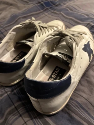 Golden goose superstar - Säljer ett par vita sneakers från Golden Goose med en ikonisk blå stjärna på sidan och blå detalj vid hälen. Skorna har snörning och är tillverkade i skinn, med en platt sula och rund tå. Perfekta för dig som gillar en avslappnad men trendig stil.