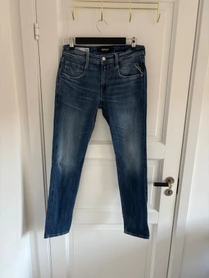 Replay Anbass blå jeans slim fit - Säljer ett par Replay Anbass jeans i klassisk blå tvätt med snygga slitningar. Modellen har rak passform, fem fickor och detaljerade sömmar på bakfickorna. Jeansen är tillverkade i mjukt bomullsmaterial och har normal midja med knapp och dragkedja framtill.