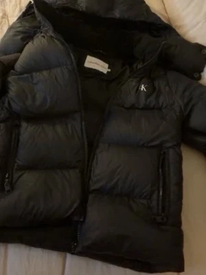Svart pufferjacka från Calvin Klein - Svart pufferjacka från Calvin Klein med diskret CK-logga på bröstet. Jackan har hög krage, dragkedja framtill och två sidofickor med dragkedja. Materialet är syntet och jackan är vadderad för extra värme. Perfekt för kalla dagar.