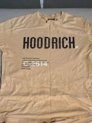 Beige HOODRICH t-shirt med tryck - Snygg beige t-shirt från HOODRICH i storlek S. Framsidan har ett stort svart HOODRICH-tryck och textdetaljer i svart och vitt. T-shirten har rund hals och är kortärmad, tillverkad i mjuk bomull som är skön mot huden. Perfekt för dig som gillar streetwear och vill sticka ut.