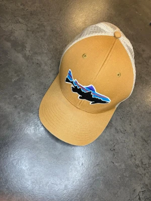 Patagonia  - Säljer en snygg truckerkeps från Patagonia i beige och brunt med mesh på baksidan och justerbar snapback. Framtill finns ett broderat fiskemärke i svart, blått och lila. Perfekt för dig som gillar friluftsliv och streetwear.