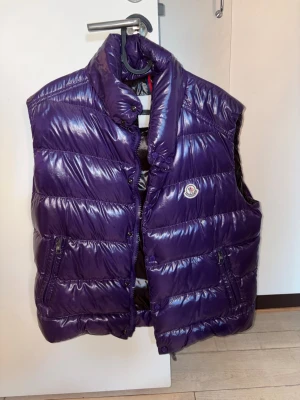Lila dunväst från Moncler - Säljer en glansig lila dunväst från Moncler med hög krage och två dragkedjefickor. Västen har quiltad design och klassisk Moncler-logga på bröstet. Insidan är brun och har en unik serietidningsetikett. Perfekt för dig som vill ha en statement piece.