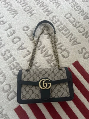 Gucci GG Supreme axelväska - Säljer en ikonisk Gucci GG Supreme axelväska med kedjerem och detaljer i svart skinn. Väskan har klassiskt GG-mönster i beige och svart, guldfärgat GG-spänne och innerfack med dragkedja. Perfekt storlek för det viktigaste och riktigt snygg design.