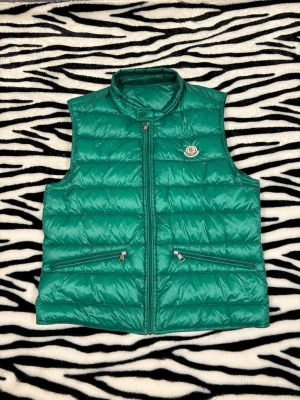 Moncler väst - Säljer en grön dunväst från Moncler med quiltad design och två dragkedjeförsedda fickor framtill. Västen har ståkrage och klassisk Moncler-logga på bröstet. Perfekt för lager-på-lager och snygg att styla till streetwear. Storlek 3 vilket är xs/s hör av dig vid minsta fundering 😌