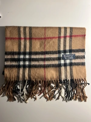 Beige rutig halsduk från Burberrys - Klassisk beige halsduk från Burberrys i 100% kashmir. Den har det ikoniska rutiga mönstret i beige, svart, vitt och rött samt fransar i ändarna. Perfekt accessoar för att lyfta din outfit med en tidlös brittisk vibe. (10C)