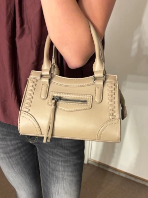 Beige handväska med snygga detaljer - Säljer en beige handväska i skinnimitation med stilrena handtag och dekorativa sömmar längs kanterna. Väskan har en liten dragkedjeficka framtill och coola metallringar vid handtagen. Perfekt storlek för det viktigaste och riktigt trendig look.