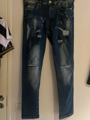 Blå jeans från Supply & Demand NYC - Säljer ett par blå jeans från Supply & Demand New York City i storlek 32W M. Jeansen har slitna detaljer, små hål och blekta partier framtill för en cool, urban look. Modellen är rak med klassiska fem fickor och tillverkad i jeansmaterial.