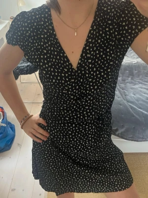 Svart blommig omlottklänning - Säljer en svart kort omlottklänning med små vita blommor. Klänningen har v-ringad hals, korta ärmar och knyt i midjan för en snygg passform. Perfekt för dig som gillar en feminin och trendig stil.