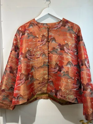 Elegant kort jacka i 100% siden med vackert orientaliskt landskapsmönster. - Elegant kort jacka i 100% siden med vackert orientaliskt landskapsmönster.  • Tillverkad av 100% mullbärssilke – mjuk, lätt och exklusiv • Konstnärligt mönster med berg och naturmotiv • Kort modell med avslappnad passform • Guldknappar ger en elegant och unik känsla • Perfekt som statement-plagg året runt  📏 Mått: Byst: 118 cm Axel: 42 cm Längd: 59 cm  🔸 Märkt storlek 4XL, men passar även M–L–XL beroende på önskad passform (oversize / relaxed fit)  Skicka gärna meddelande om du vill ha fler bild