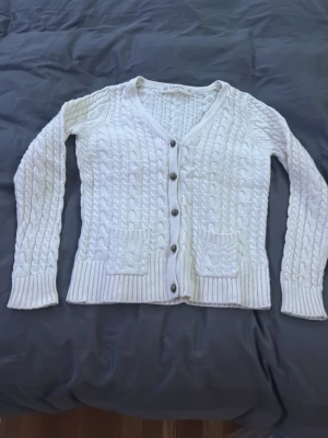 Vit kabelstickad kofta från ZARAKNIT - Supermysig vit kabelstickad kofta från ZARAKNIT med v-ringning och knappar framtill. Koftan har två fickor och ribbade muddar vid ärmslut och nederkant. Materialmix av bomull, viskos, nylon och angora. Köpt för ett tag sen men har aldrig använts 