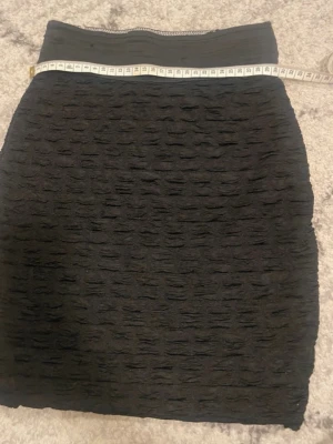 Svart kortkjol med struktur - Säljer en svart kortkjol med coolt strukturerat tyg och bred resår i midjan. Oklar i storlek men skulle säga xs-S. Kjolen har en tight passform och är perfekt om du gillar en enkel men ändå unik look. Materialet känns mjukt och stretchigt, vilket gör den bekväm att bära hela dagen.
