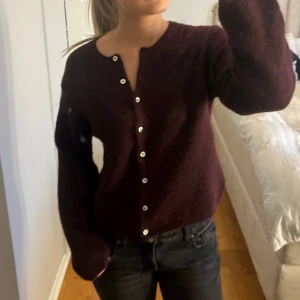 Vinröd kofta med knappar - Mysig vinröd kofta med lång ärm och rund hals. Koftan har en rak passform. Tröjan ör i alpaca blend med 30% alpaca.