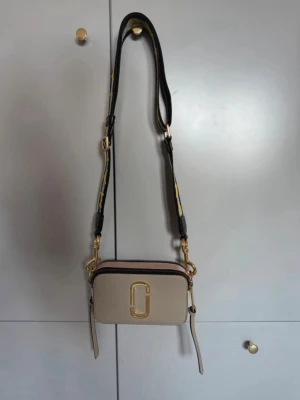 Marc Jacobs beige axelväska  - Beige axelväska från Marc Jacobs med guldfärgade detaljer och logotyp framtill. Väskan har en bred, justerbar axelrem med svart, grå och gul text. Två fack med dragkedja och en ficka på baksidan. Sparsamt använd. Lite nagg på märket på framsidan, se bild.